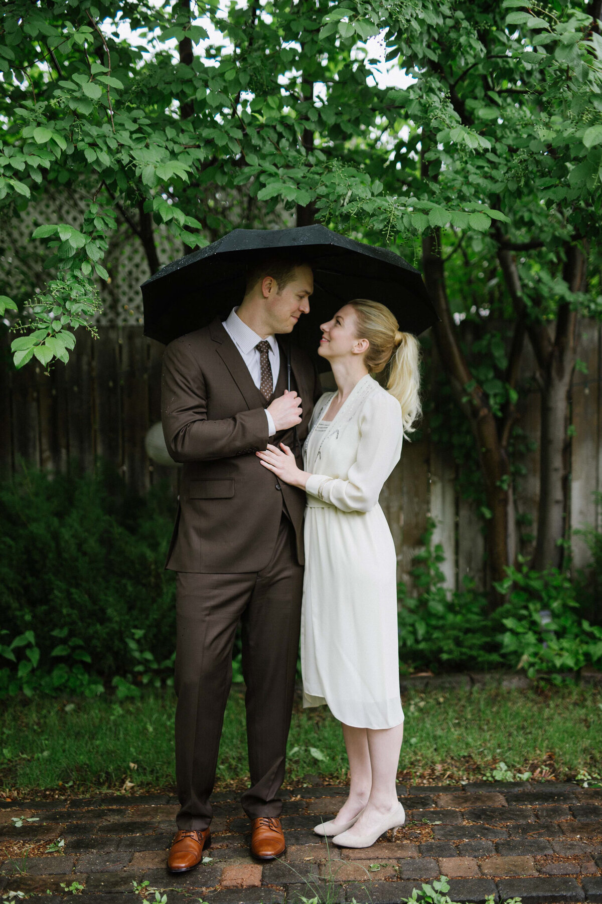 Vintage wedding inspiration, bride wearing antique knee length wedding outfit and heels, groom in a classic brown suit, couple holding umbrella against a tree backdrop, captured by Christy D. Swanberg Photography, editorial elopement and wedding photographer in Calgary, Alberta, featured on the Bronte Bride Vendor Guide.