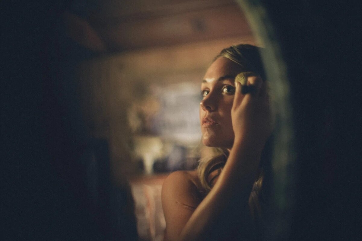 A bride is framed through a blurred foreground, applying makeup in a softly lit room, capturing a moment of bridal preparation.