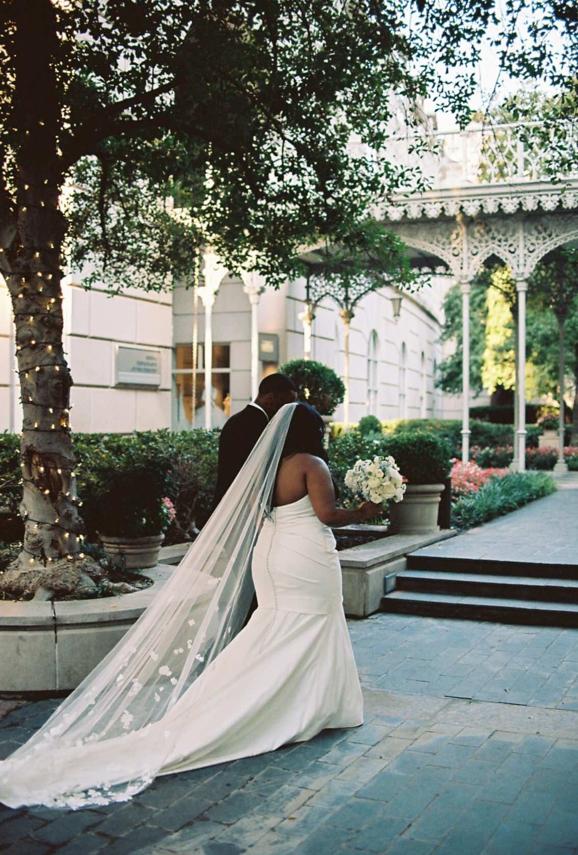 Elegant couple walking through a garden courtyard, the bride in a flowing white dress and veil, holding a bouquet. The scene is serene and romantic.