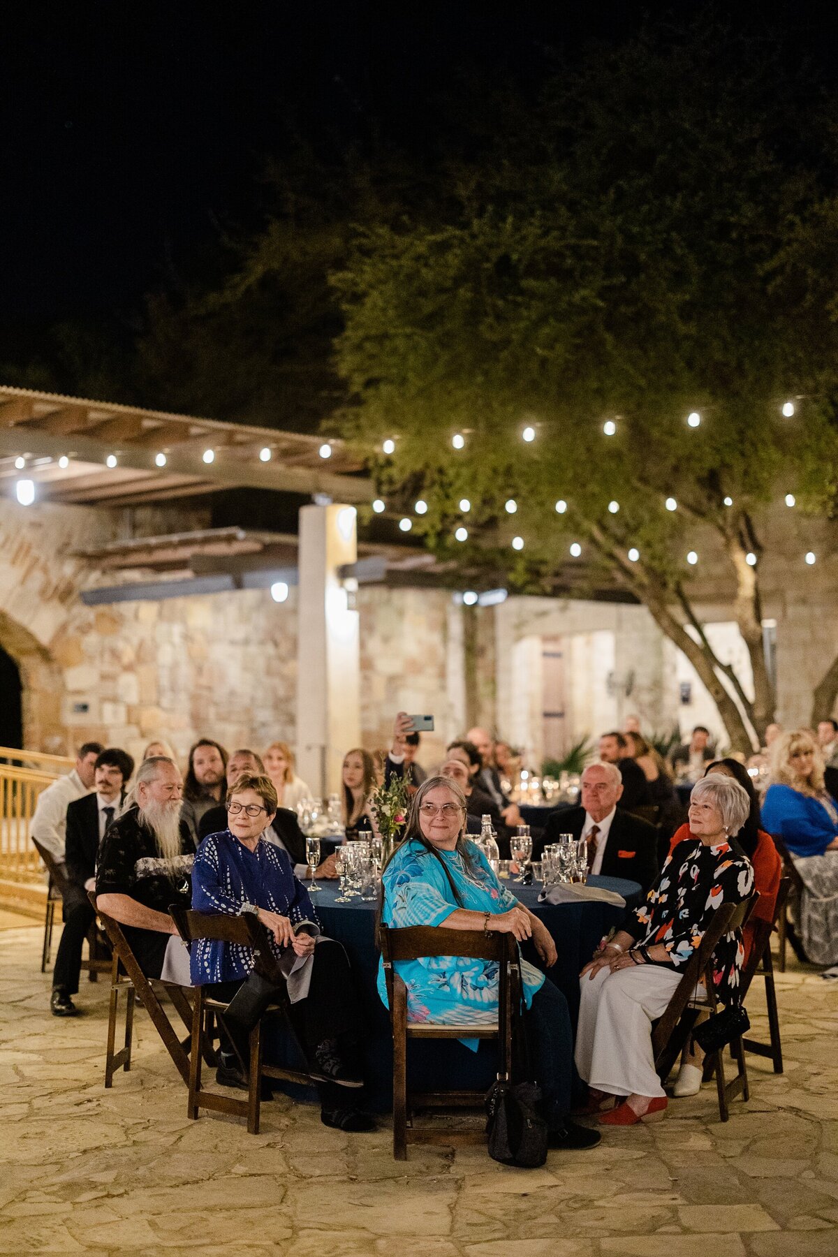 A shot of wedding guests sitting at their reception tables and watching the bride and groom during their first dance during their wedding reception at the Lady Bird Johnson Wildflower Center in Austin, Texas. The guests are all captivated by what's happening out of frame and the scene is framed by many rows of cafe lights criss crossing above them.