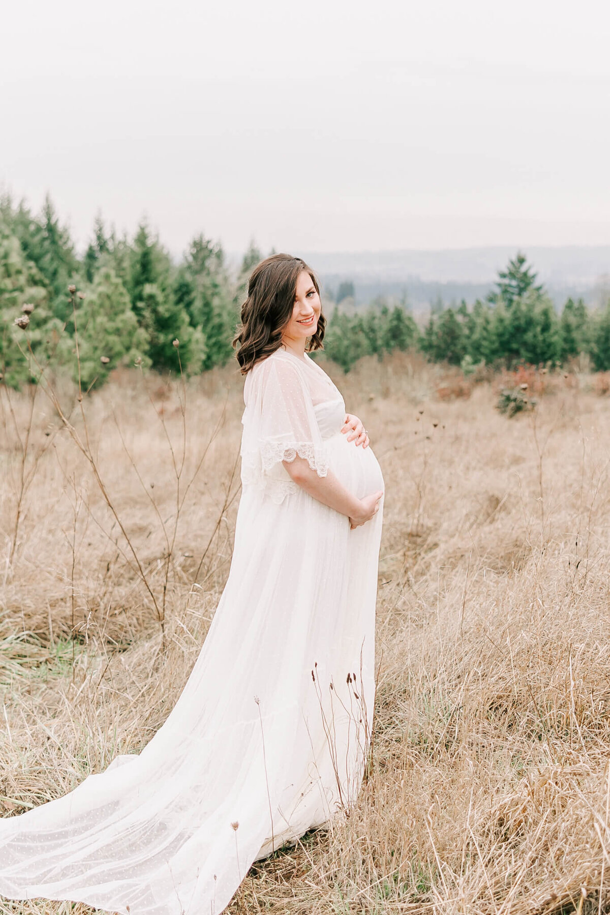 mom with hands on belly wearing white dress smiling looking at the camera for her maternity portraits in beaverton oregon