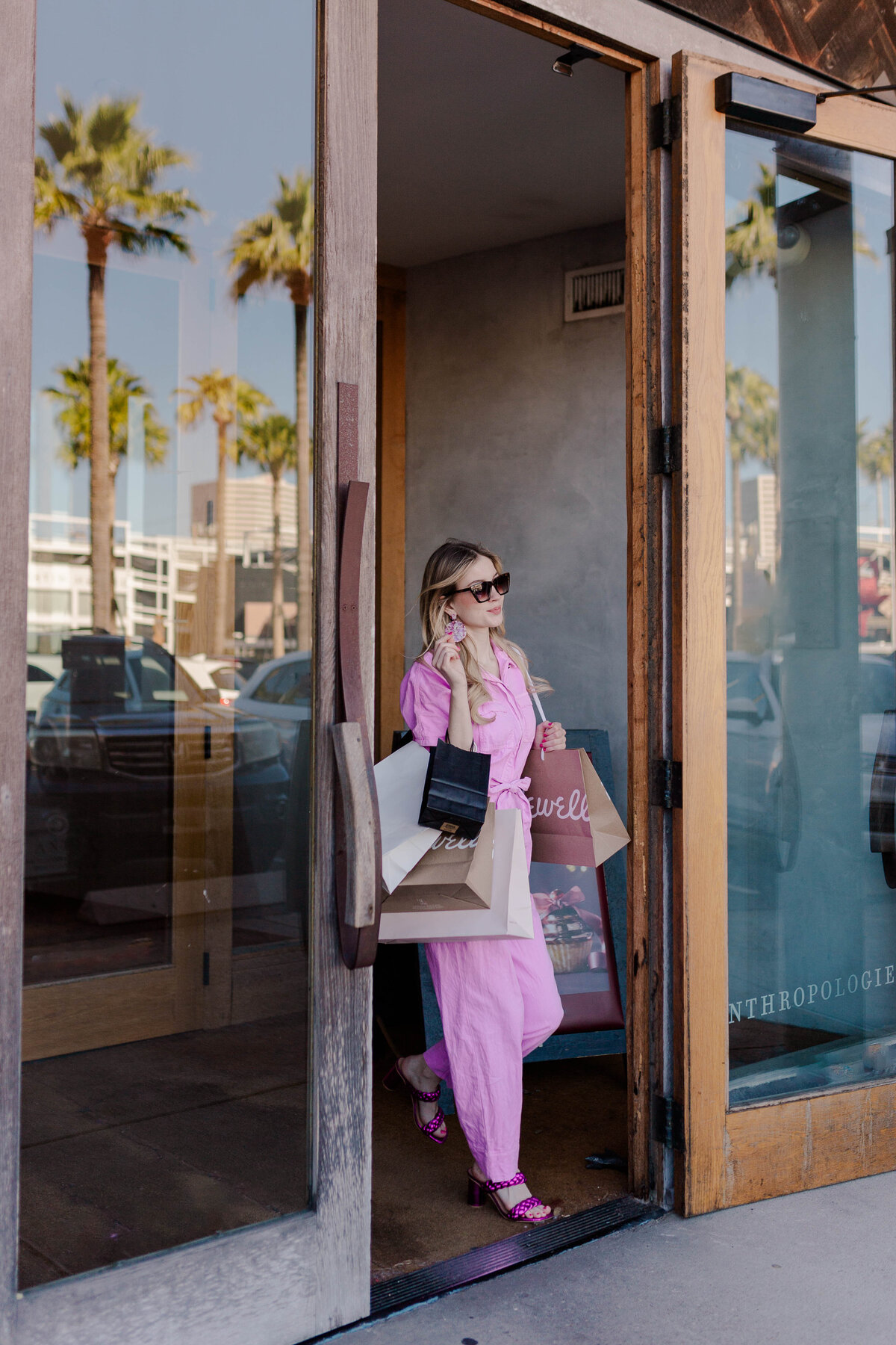 Barbie model wearing a pink jumpsuit walks out of anthropologie with shopping bags while reaching for her sunglasses
