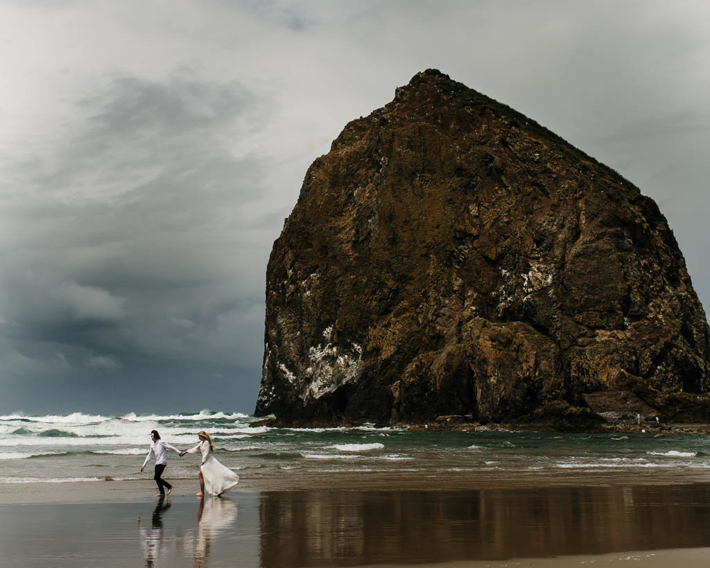 Wedding couple runs holding hands on beach  at haystack rock in Oregon