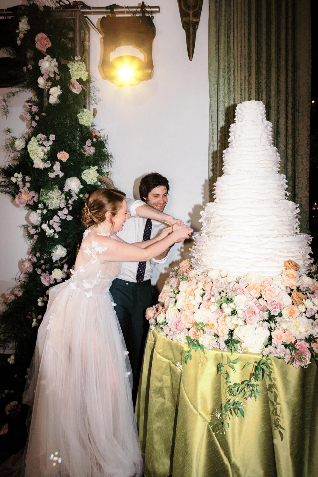 The bride and the groom are both cutting a huge white, six-layer wedding cake, with many flowers at the bottom.