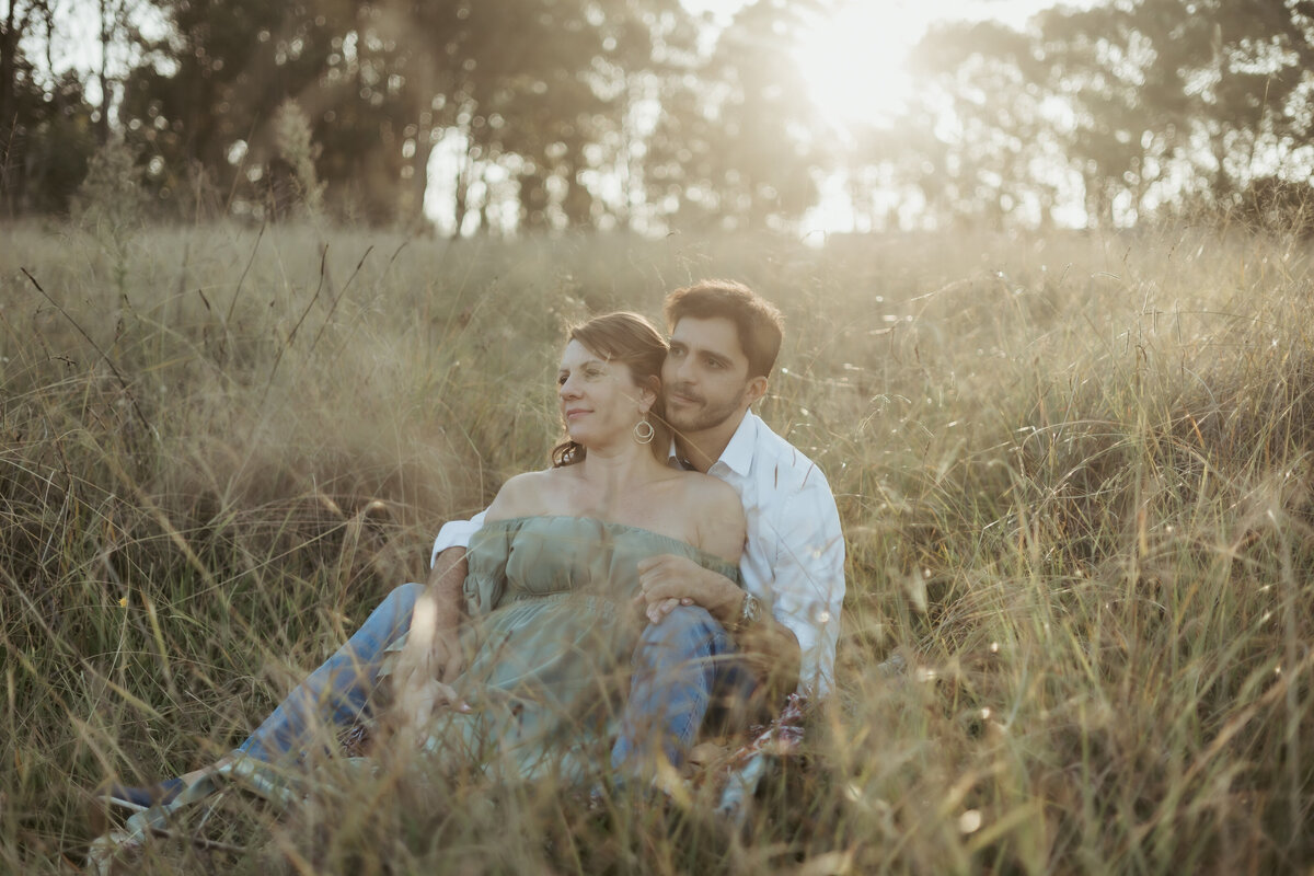 Husband and pregnant wife relaxing in a grassy field to celebrate they're pregnancy while the sun glows behind them