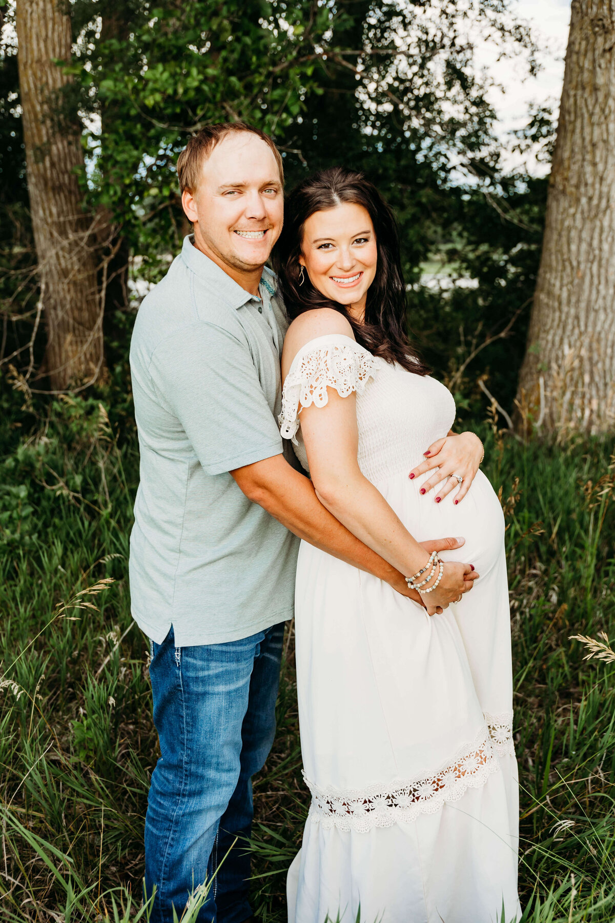 Mother to be and dad hug in front of a tree area.