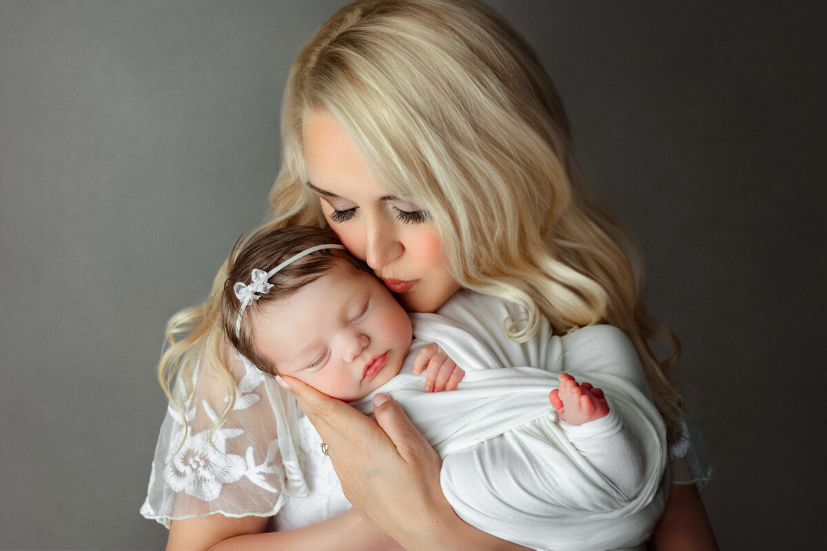 new mother wearing a white dress holding and kissing her newborn baby at a baby photography session in leesburg va