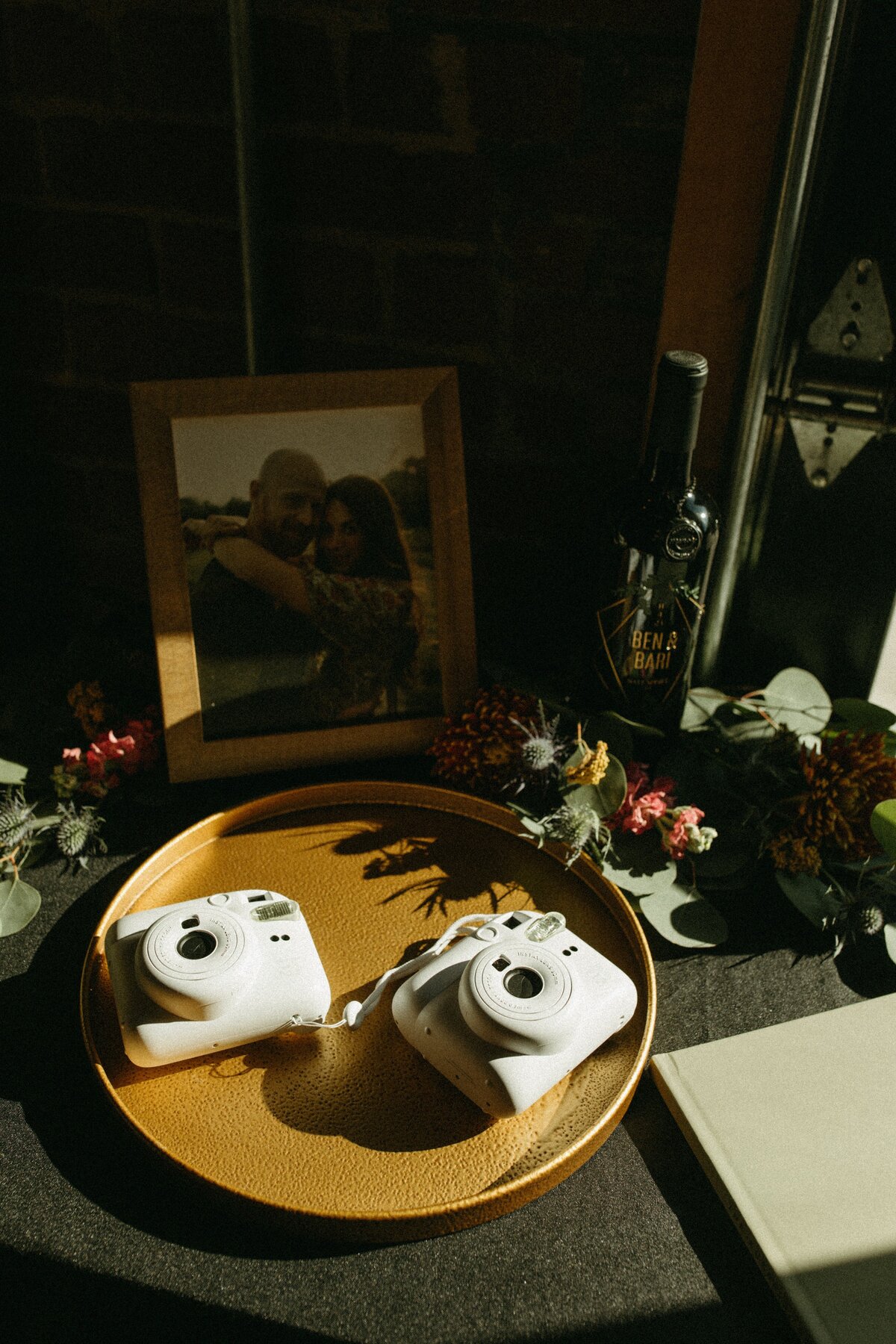 A framed couple's portrait next to two white instant cameras on a tray, with a bottle and floral decorations around, all under natural light at a Park Farm Winery wedding.