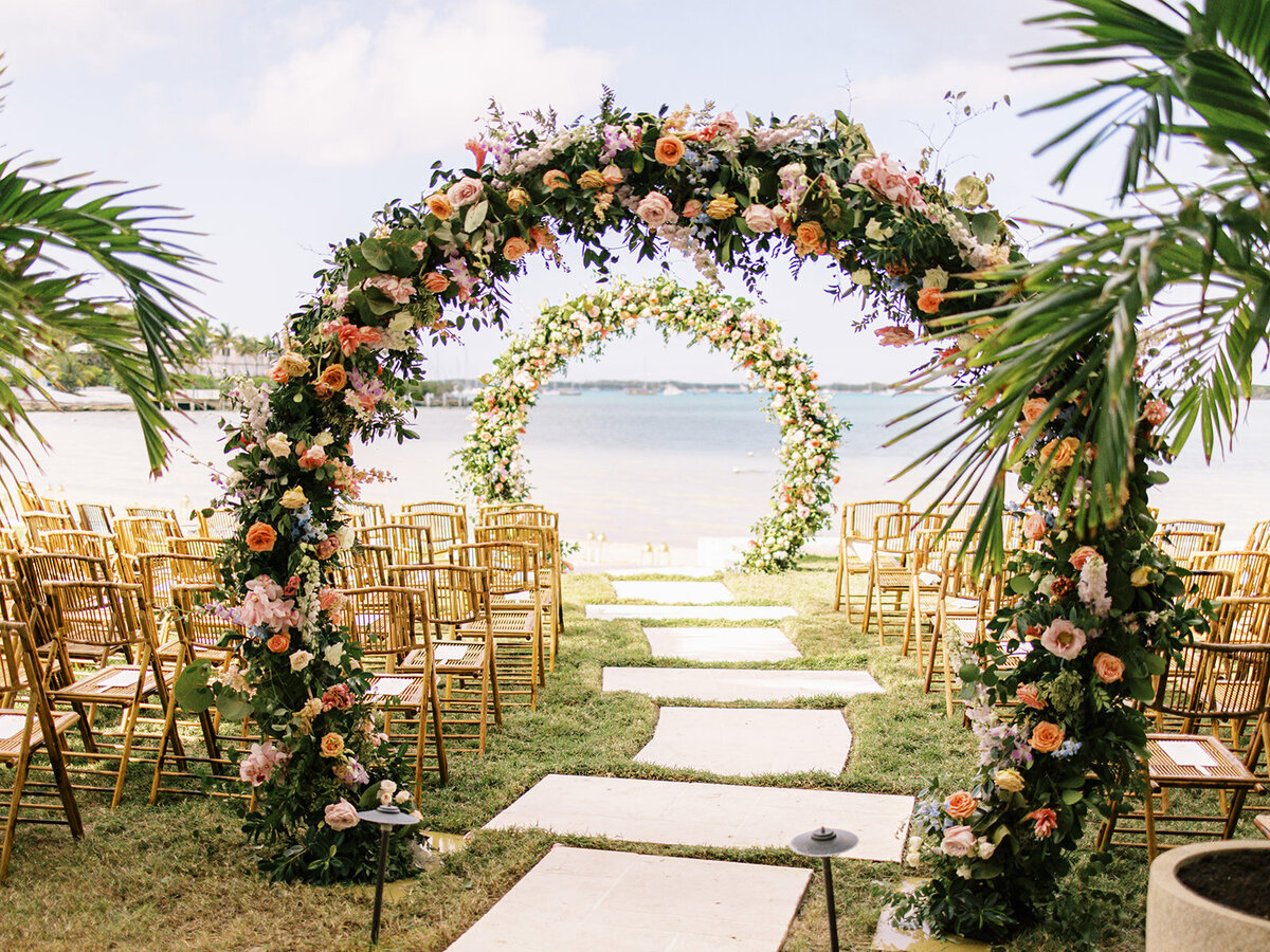 Double circle arch floral ceremony arch for spring tropical wedding ceremony. Exuma, Bahamas beach private estate destination beach wedding. Roses, tropical flowers, tropical foliage in colors of peach, dusty pink, orange, dusty blue, cream, lavender, and natural greenery. Floral Design by Rosemary & Finch Floral Design.
