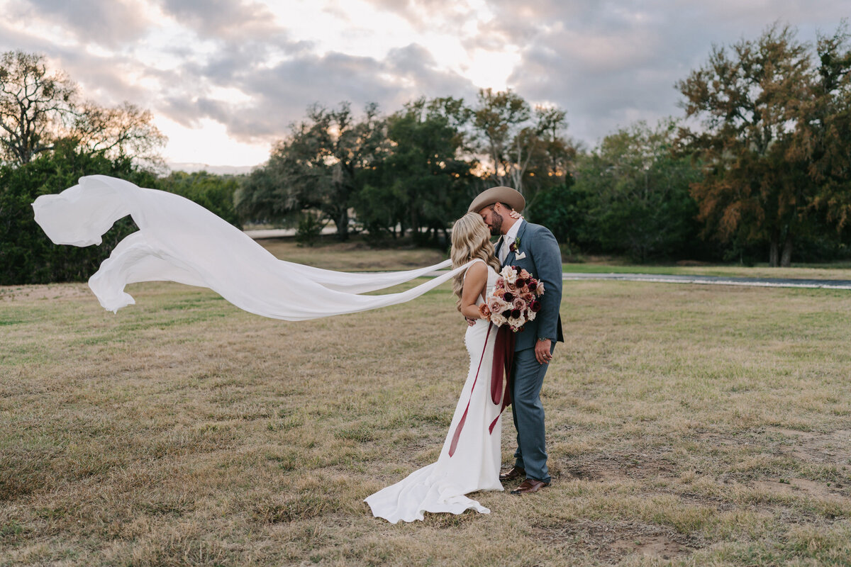 A photograph in color of Carly and Bradley on their wedding day at Morgan Creek Barn in Dripping Springs, near Austin, Texas. The bride and groom are standing on the middle of a field of grass, surrounded by oak trees, embraced in a kiss. The clouds in the sky behind them are colorful from the sunset. Her white wedding dress has wings from each shoulder that are flowing in the air to the side of her from the wind. She is holding a colorful bouquet that includes pink, white, beige, and maroon florals with maroon ribbon falling to the ground.  Wedding photography by Stacie McChesney/Vitae Weddings.