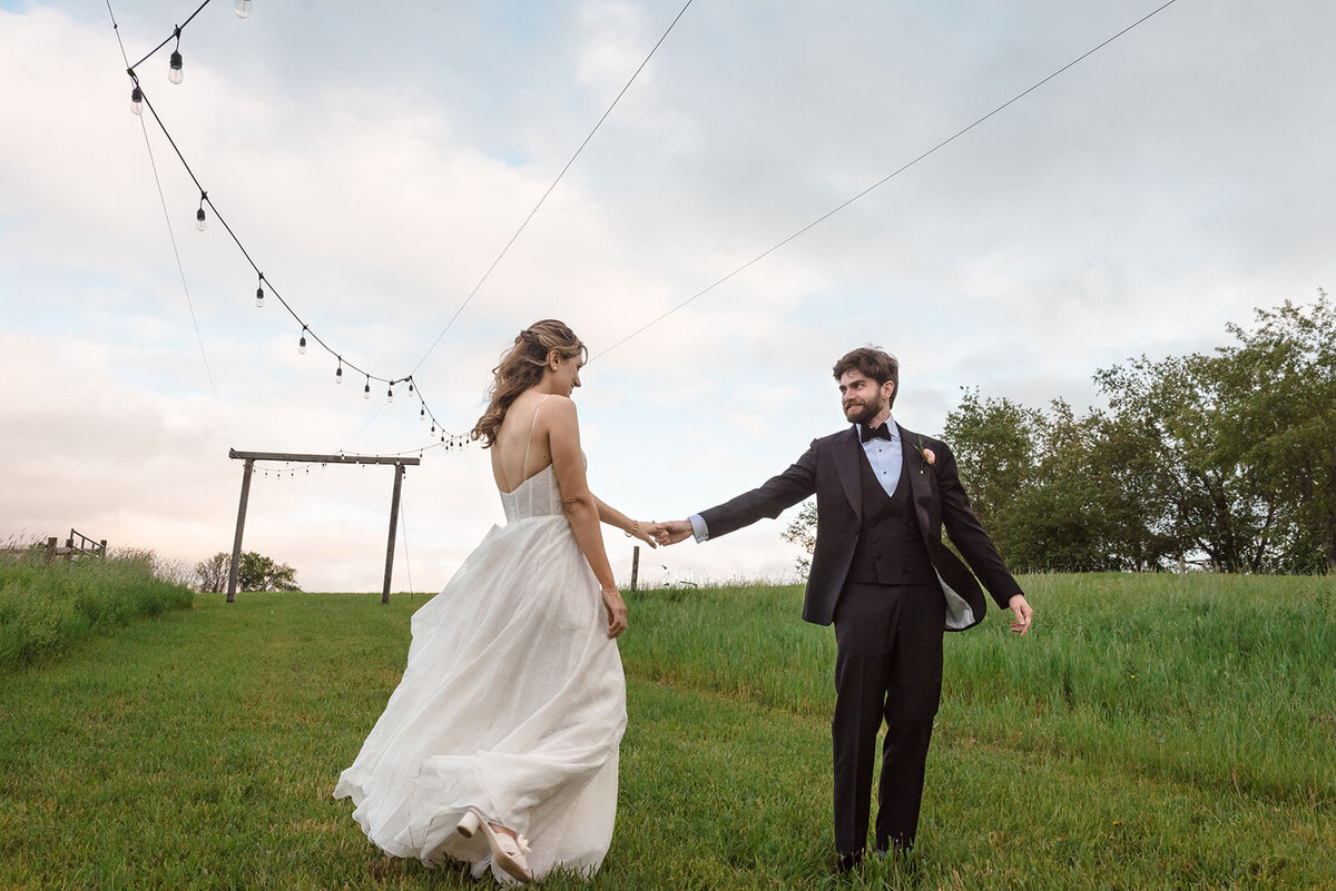 A bride and groom holding hands and walking under a sky with string lights, with open fields in the background.