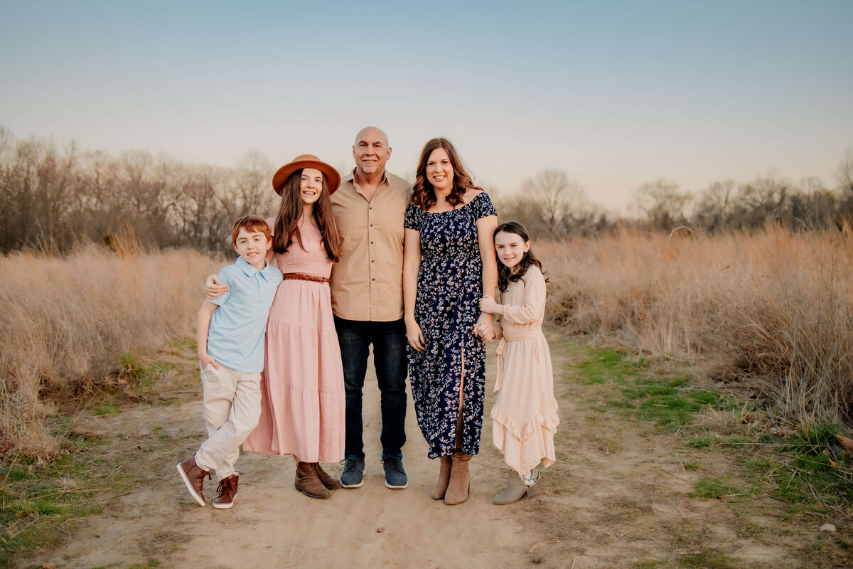 family of 5 standing side by side looking at the camera  on a dirt path  between a field