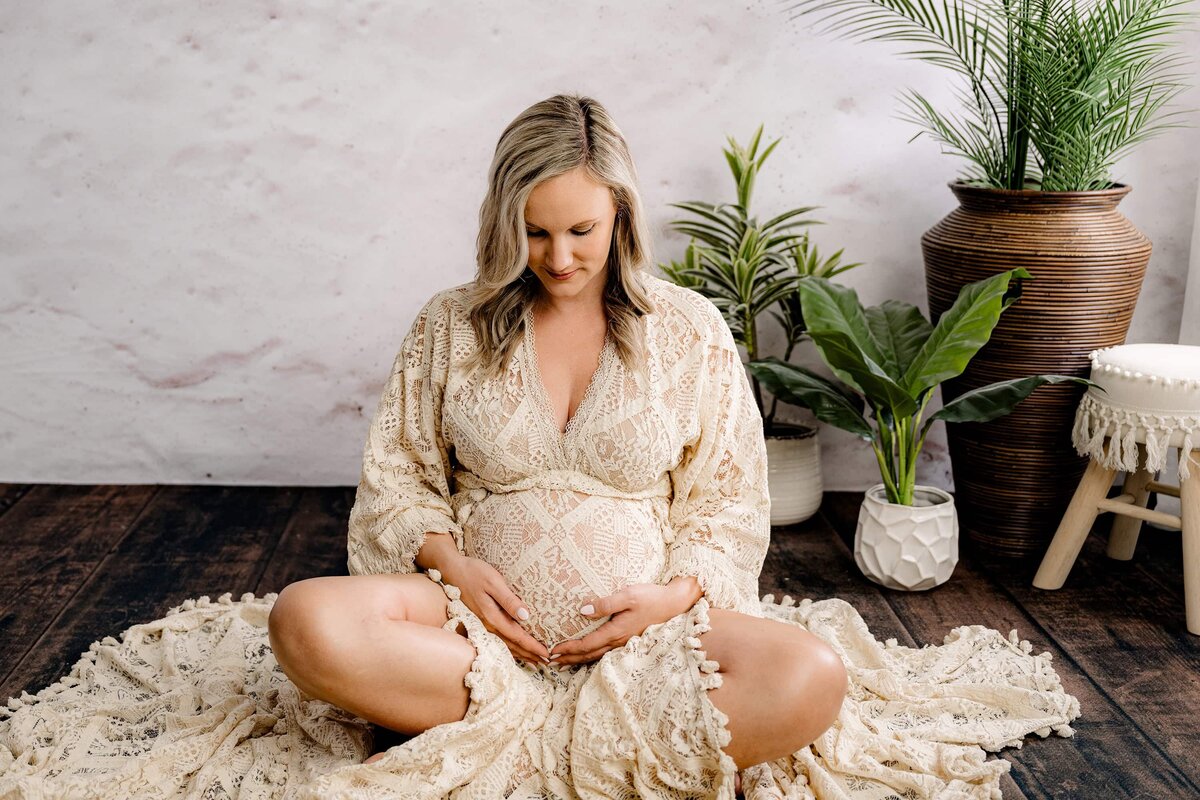 Charlotte Area expecting mother sits in the floor in a boho style dress cross-legged looking down towards her belly with both hands on her belly