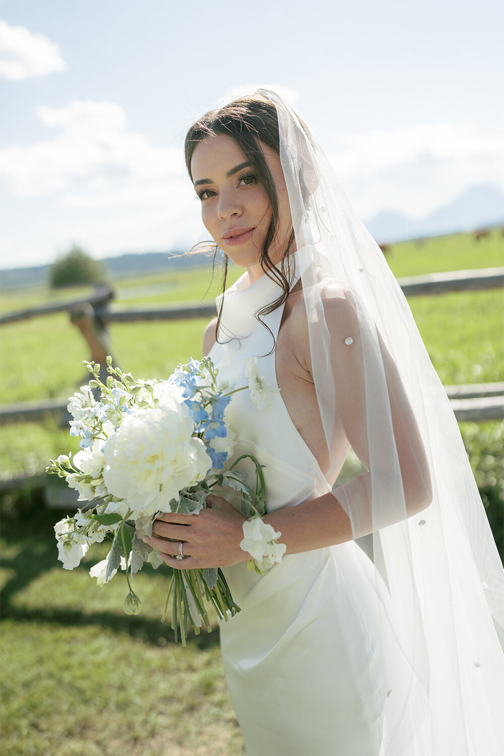 Bridal portrait with veil blowing in the wind, holding a blue and white bridal bouquet, against the backdrop of the Teton Mountains during a French blue summer wedding in Jackson, Wyoming..