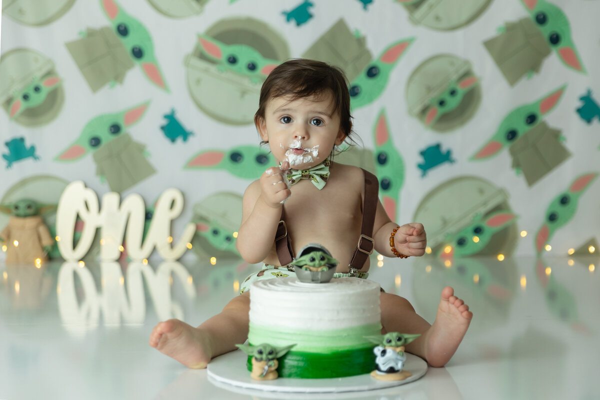 A toddler boy in a green bowtie and brown suspenders eats a baby yoda themed cake for his first birthday