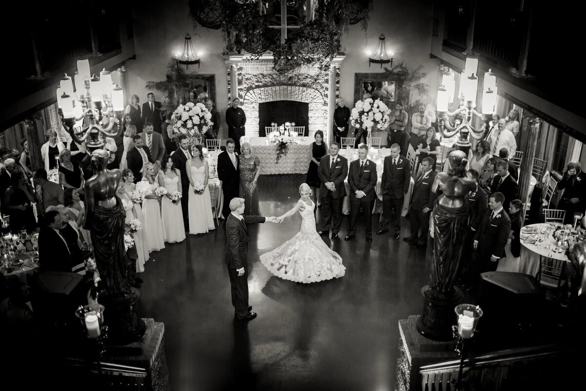 High-angle view of a bride and groom during their first dance with guests encircling them, in an elegant venue with a chandelier.