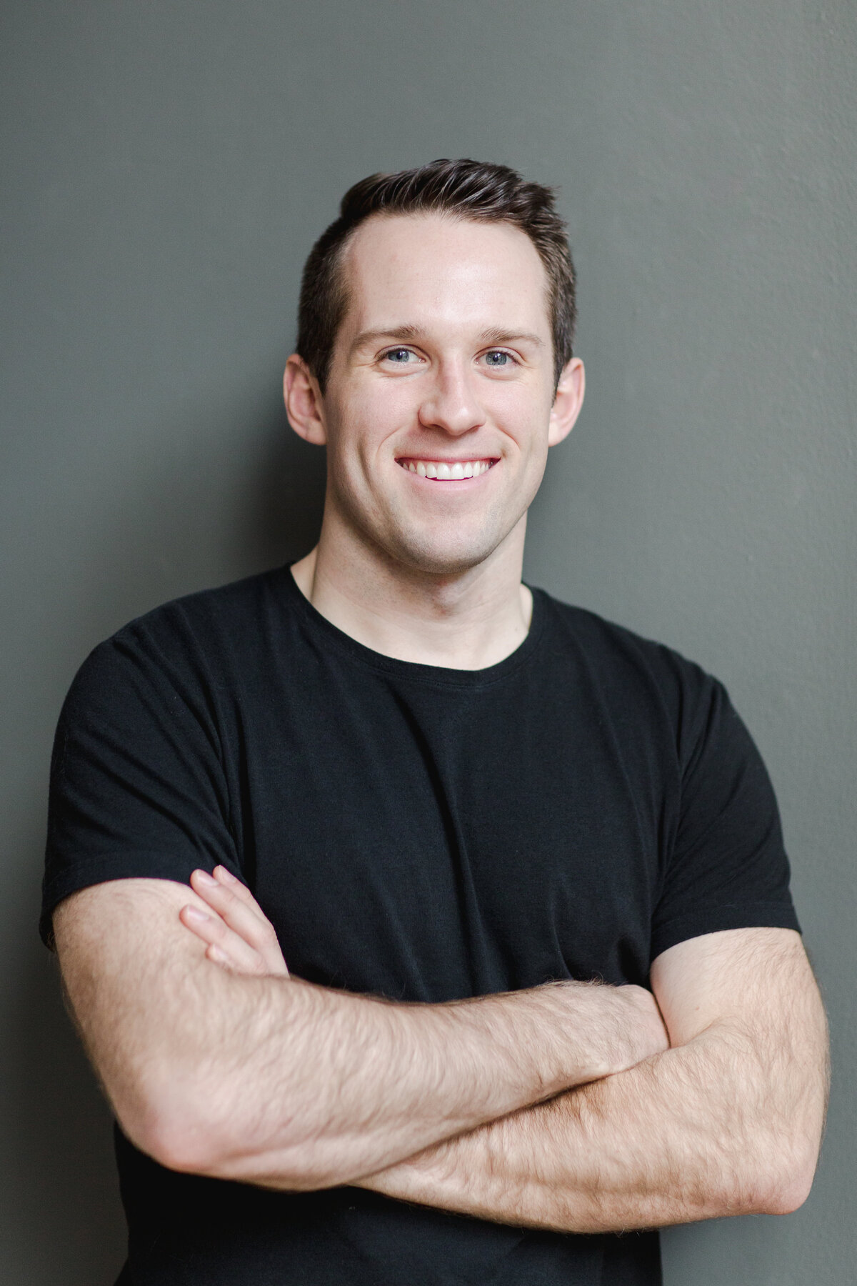 man in black top with his arms folded and leaning up against a wall smiling at the camera