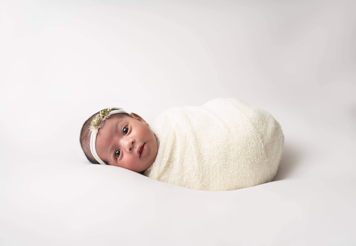 A newborn baby girl swaddled in a cream blanket stares at the camera during her session with an Asheville Newborn Photographer
