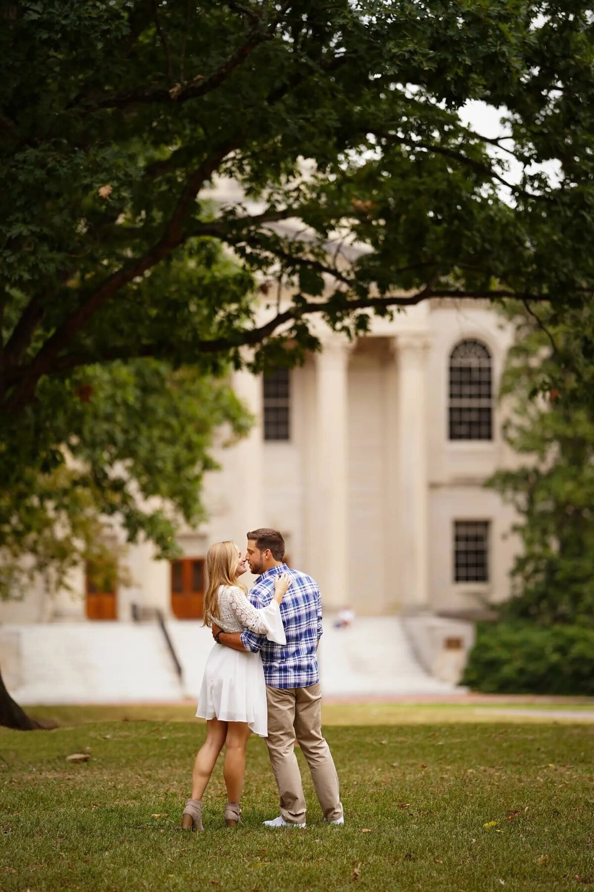 A couple embracing in front of a grand building with large columns, surrounded by lush trees