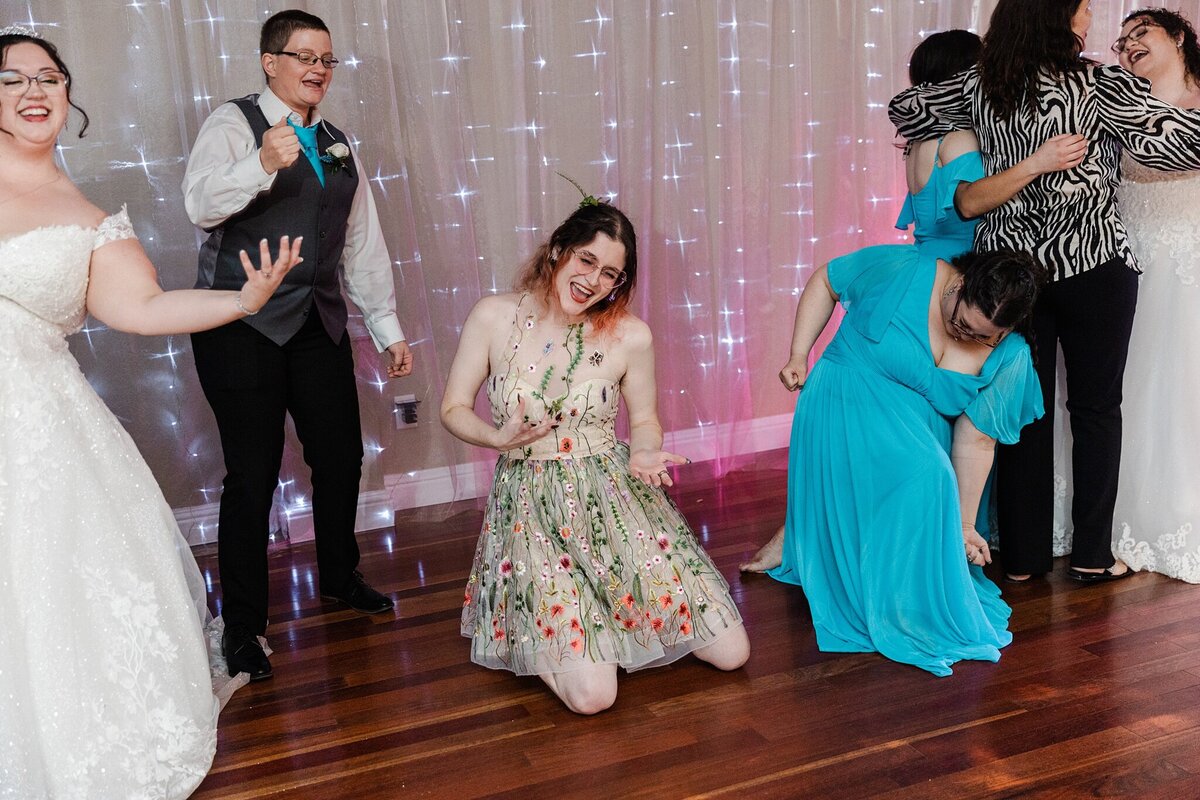 A bride and multiple wedding guests playing air guitar and rocking out during a wedding reception at the A and M Gardens in Azle, Texas. The bride is on the far left and is wearing a long, flowing, intricate, white dress. The other bride can be seen talking to guests on the far right.