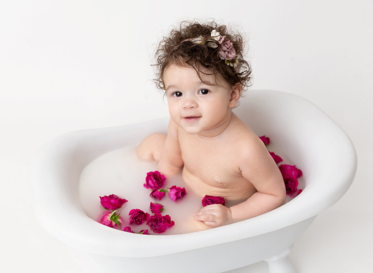 Baby girl sitting in milk bath for first birthday photoshoot. There are fresh flower petals in the bath. Baby is smiling at the camera.