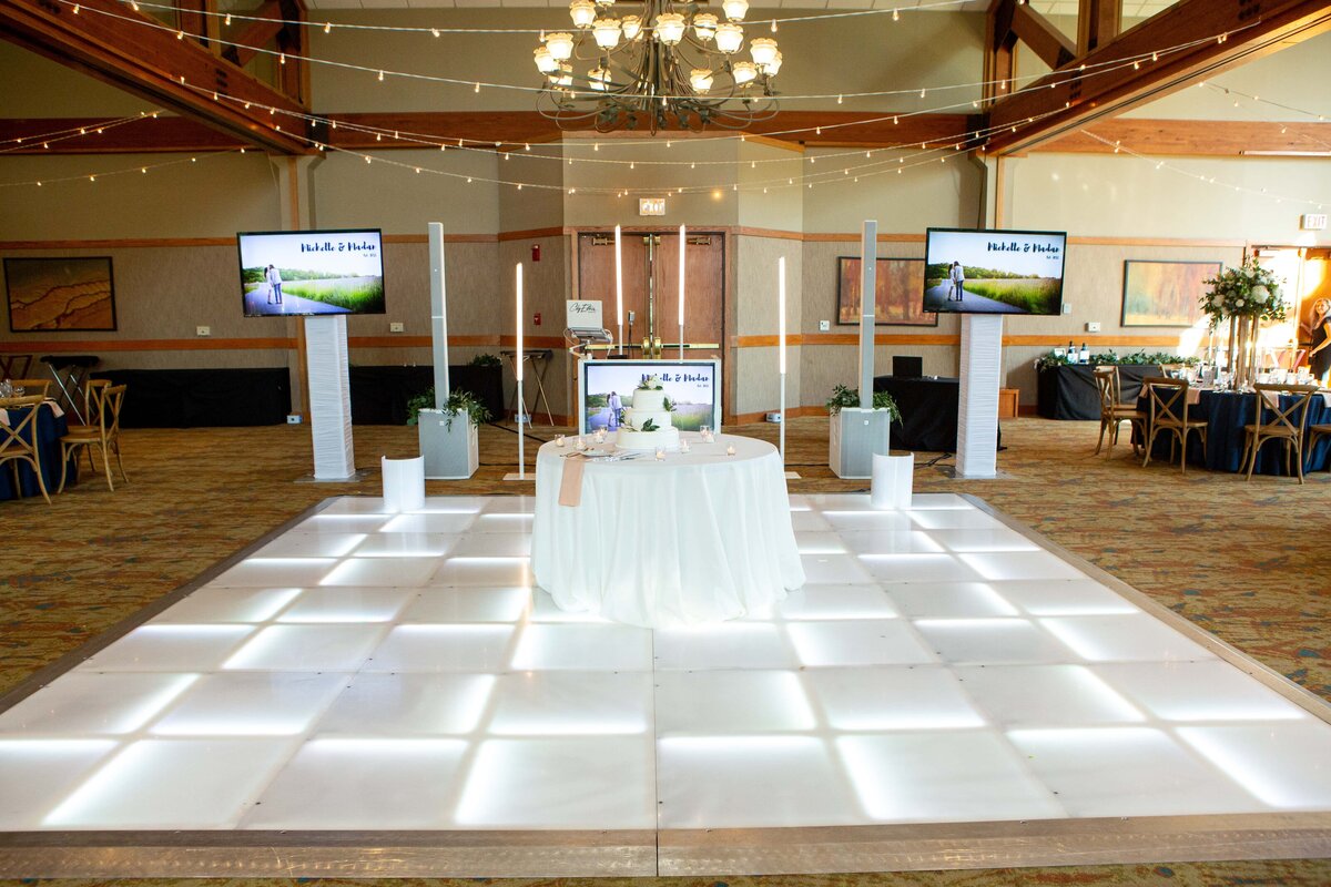 An event hall with a dance floor lit by square lights, surrounded by tables covered with white cloths and decorative plants, and monitors displaying coastal images, ideal for park farm winery weddings.