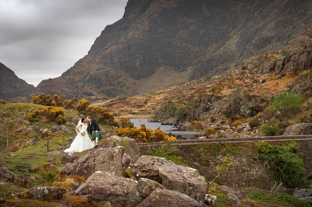 Bride in A-line wedding dress standing on an old stone bridge with her groom wearing a Scottish kilt at the Gap of Dunloe