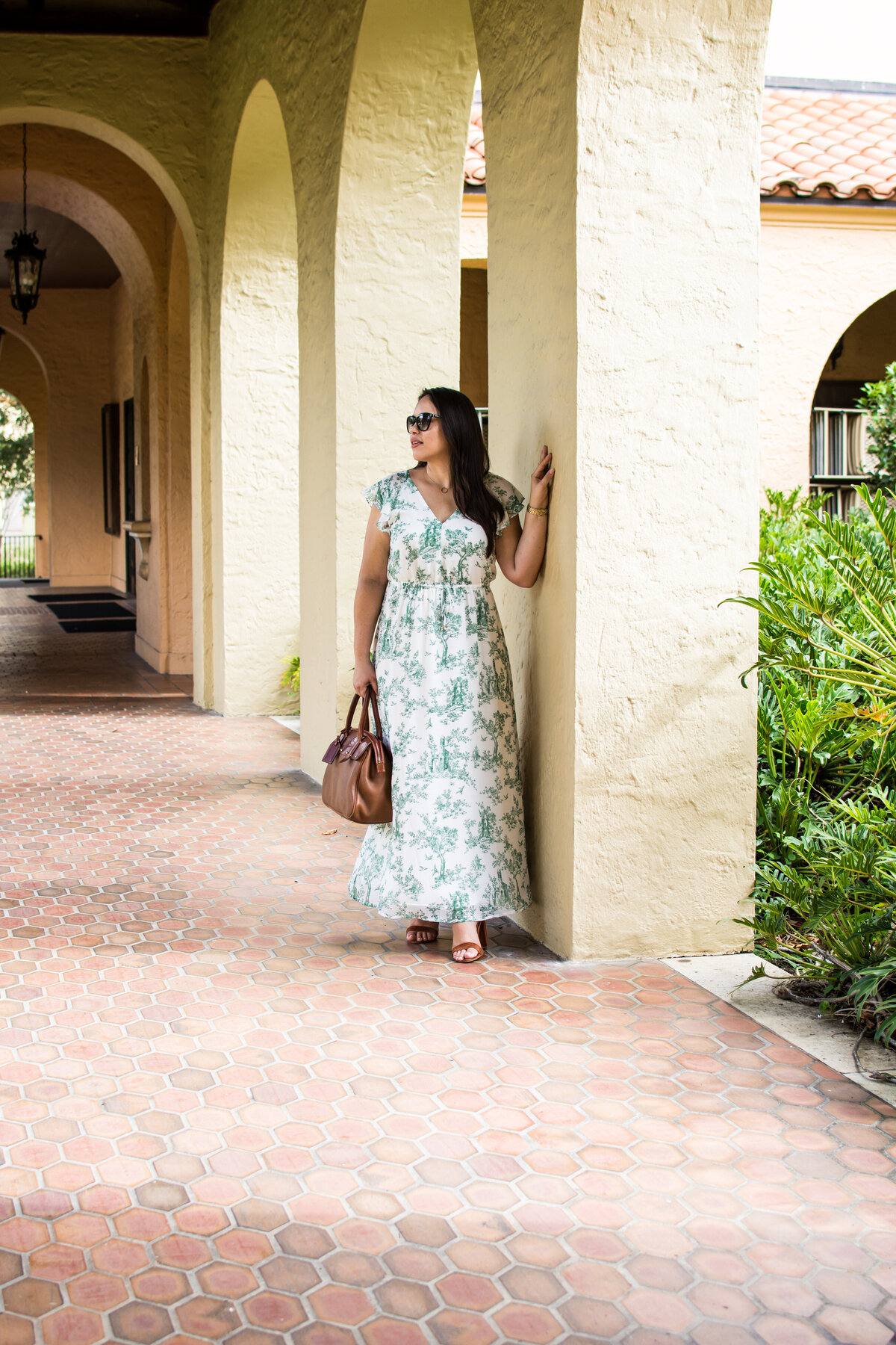 Woman with dark hair and green floral dress stands in covered walkway with arches