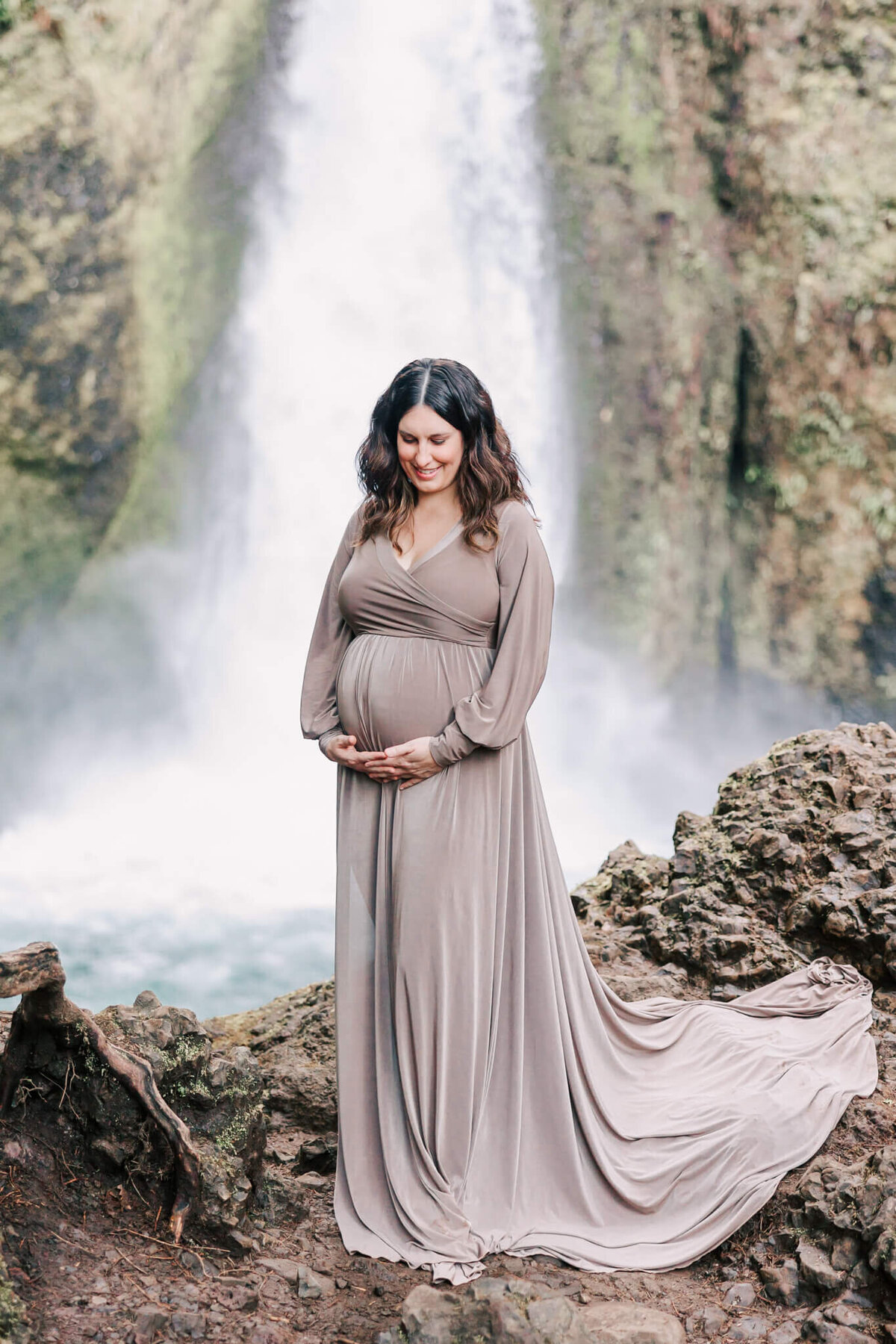 pregnant woman holding belly standing in front of wahclella falls in the columbia river gorge in oregon. she is wearing a tan dress.