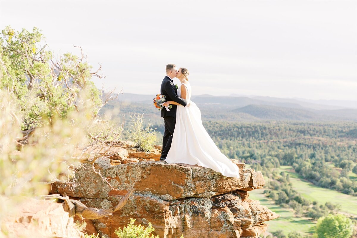 A bride and groom stand on a rocky cliff overlooking a scenic valley, embodying the romance and adventure of a luxury destination wedding experience.