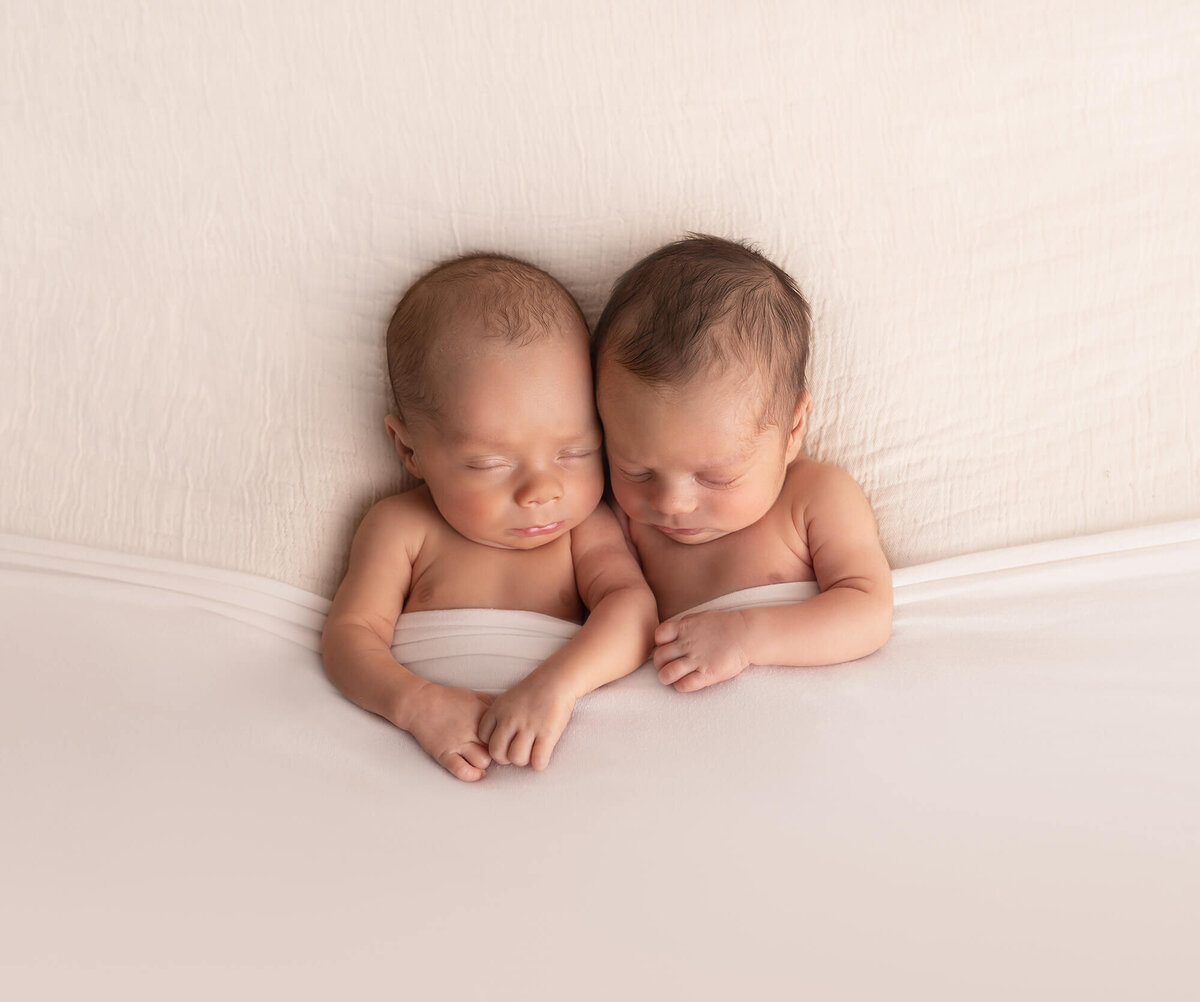 Newborn twin boys cuddle under a white blanket