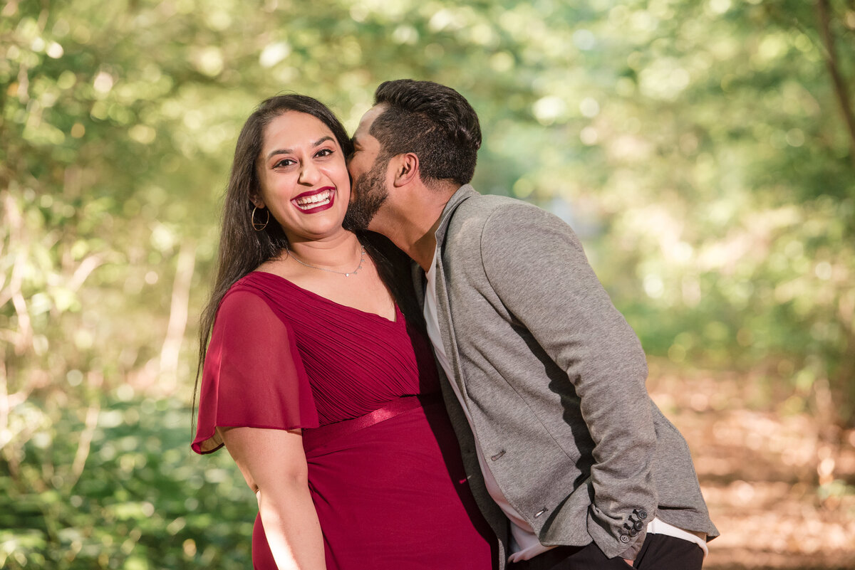 Indian-couple-laughing-and-giggling-as-the-man-in-a-grey-suit-jacket-kisses-the-woman-in-a-red-dress-on-her-cheek-amid-greenery-at-Jetton-Park