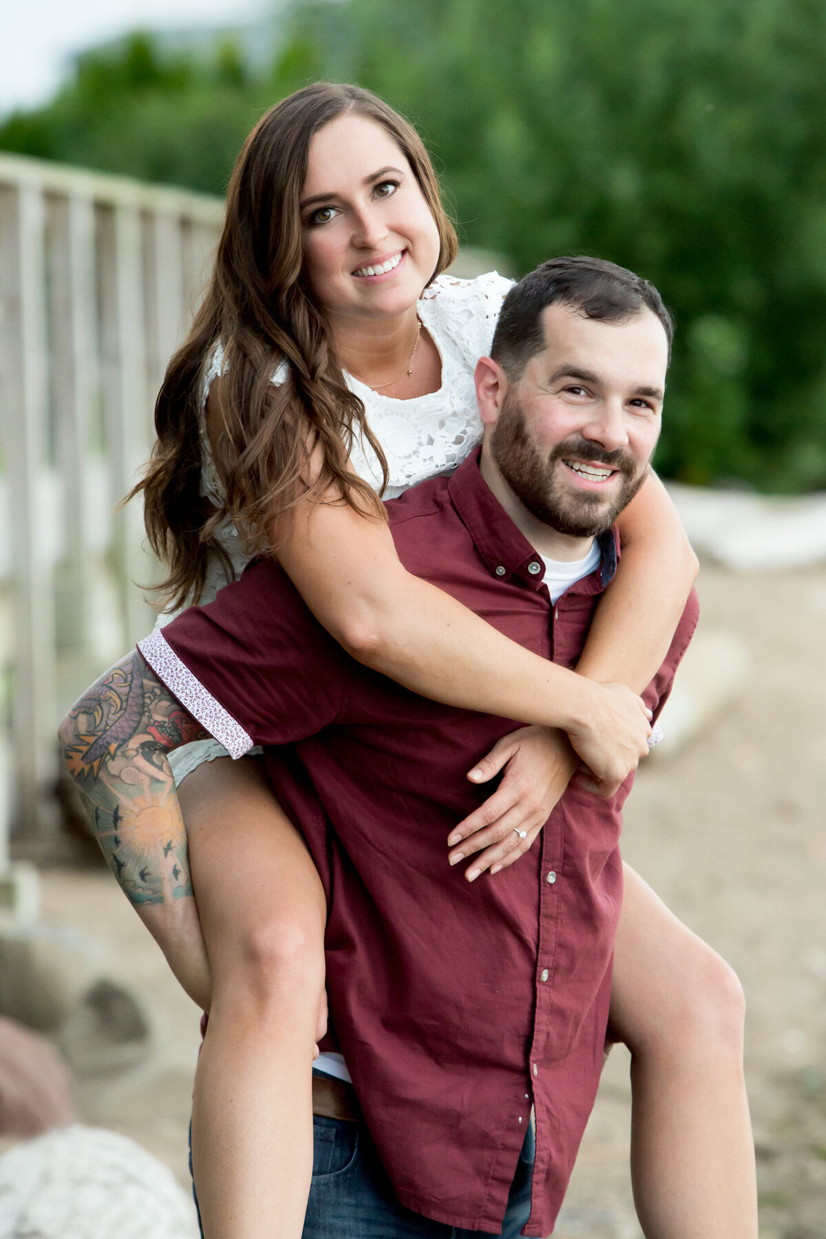 A man in a dark red shirt gives a piggyback ride to his fiance with long brown hair in a white dress Piermont engagement session New York wedding photographer