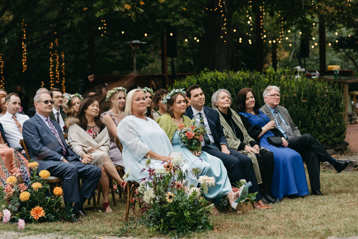 Wedding guests seated at an outdoor ceremony lined with abundant floral aisle arrangements featuring dahlias, roses, cosmos, and garden greenery, creating an immersive whimsical garden aesthetic by a Northwest Arkansas wedding florist.