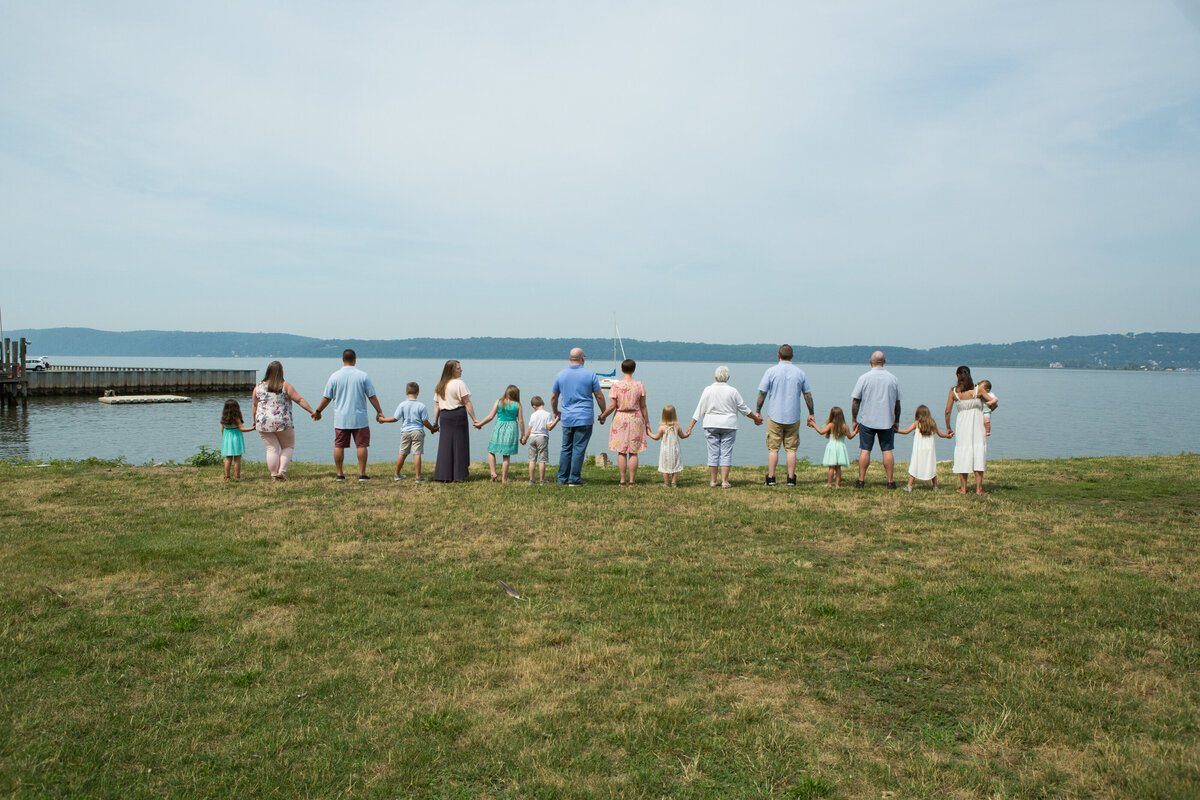 An extended family of 15 people holds hands in front of the Hudson River Westchester family photographer Milestone Images