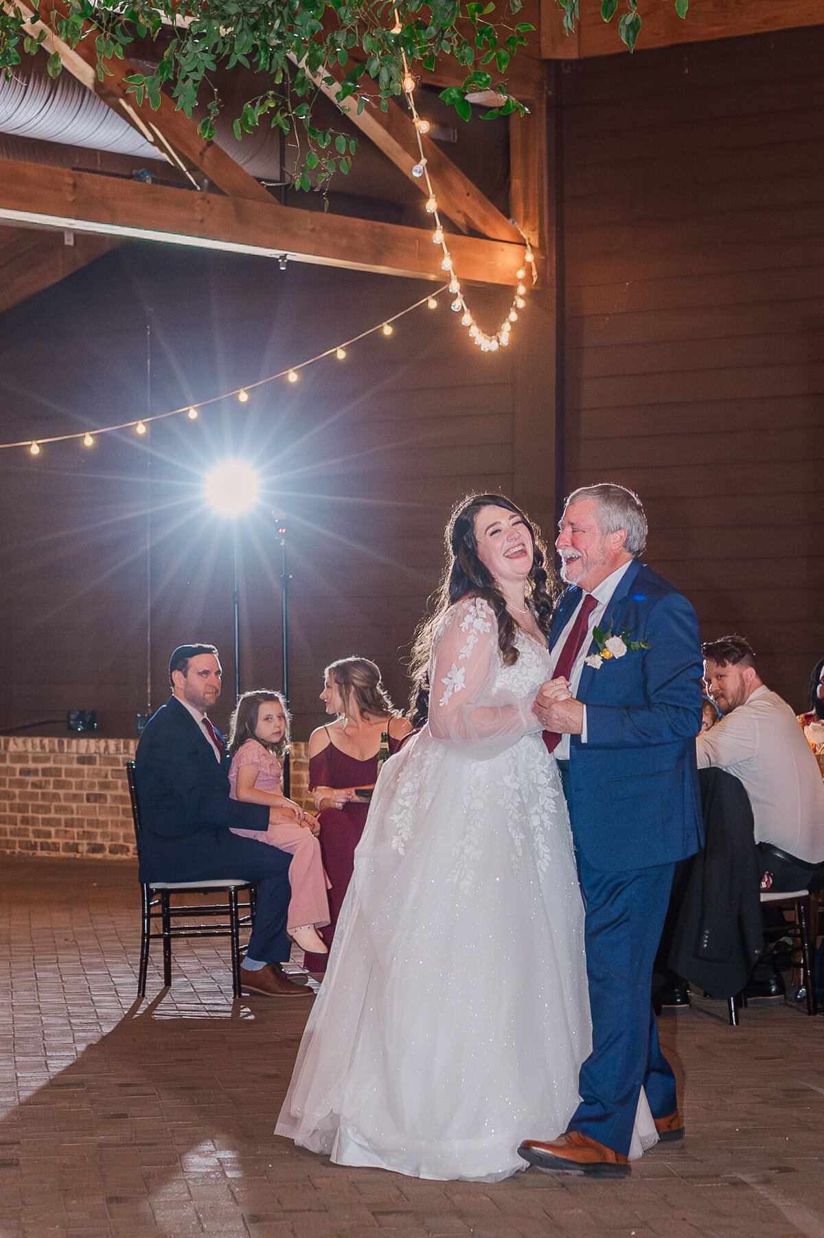 A laughing bride dancing with her father at her wedding reception underneath warm lighting at The Sutherland by JoLynn Photography, a Raleigh wedding photographer