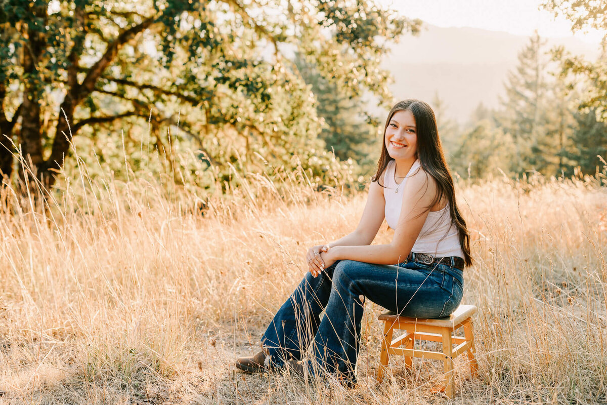 A high school senior girl poses for senior portraits at Fitton Green Corvallis, OR. She's sitting on a stool looking at the camera and smiling. Jen White Photography Albany, OR