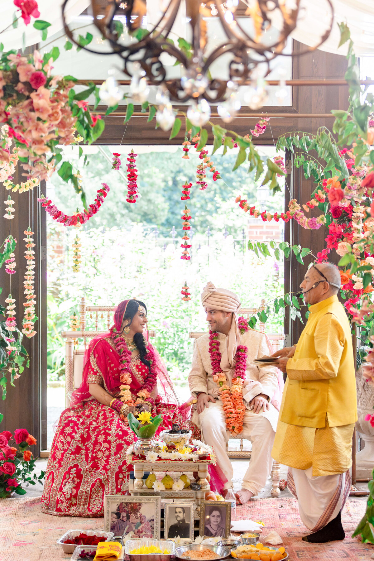 A couple in traditional Indian attire sit facing each other at a wedding ceremony. The setting is adorned with colorful flowers and garlands, and an officiant stands nearby. The room is decorated with intricate floral arrangements and offerings.