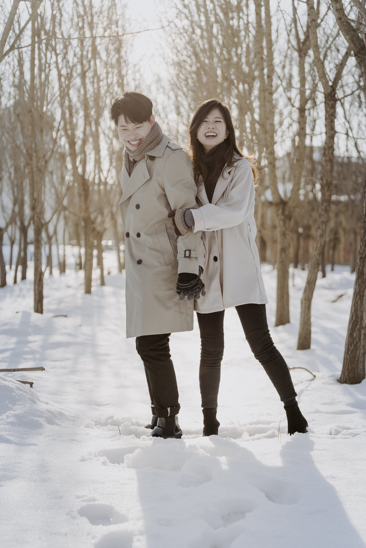 the couple laughing and the bride wears beige long coat and black turtke neck and black pants while the groom wears brown long coat and black pants with brown scarf
