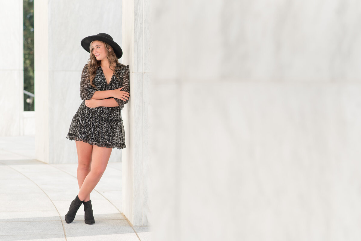 Senior girl wearing trendy black hat leaning against white wall at Founder's Hall in Hershey, Pennsylvania looking right with her feet crossed.