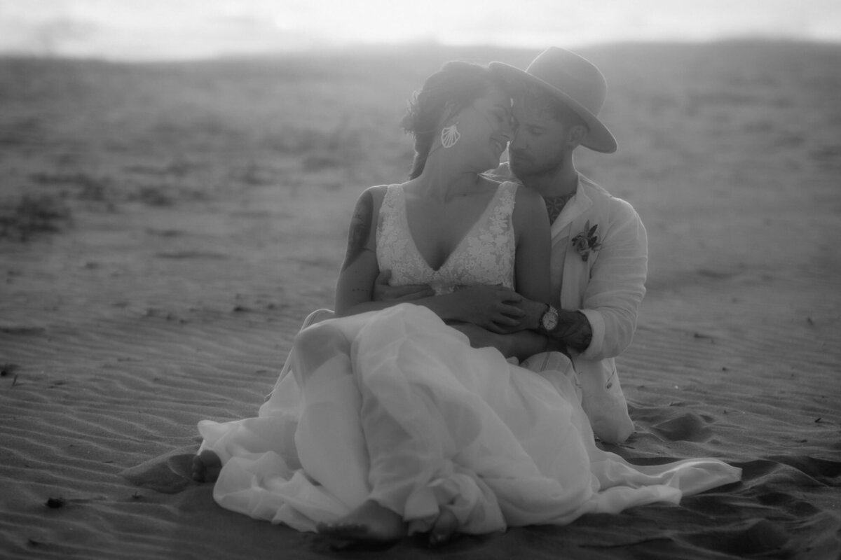 Ein romantischer Moment, als die Braut sich im Sand sitzend rücklinks an ihren Mann lehnt.