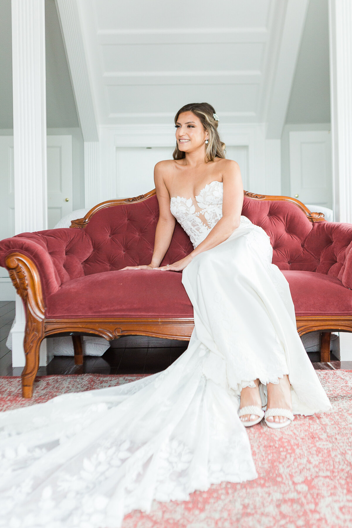 Bride is sitting on a burgundy velvet touch for a formal bridal portrait. She is smiling over her shoulder in the distance. Captured by best Massachusetts wedding photographer Lia Rose Weddings.