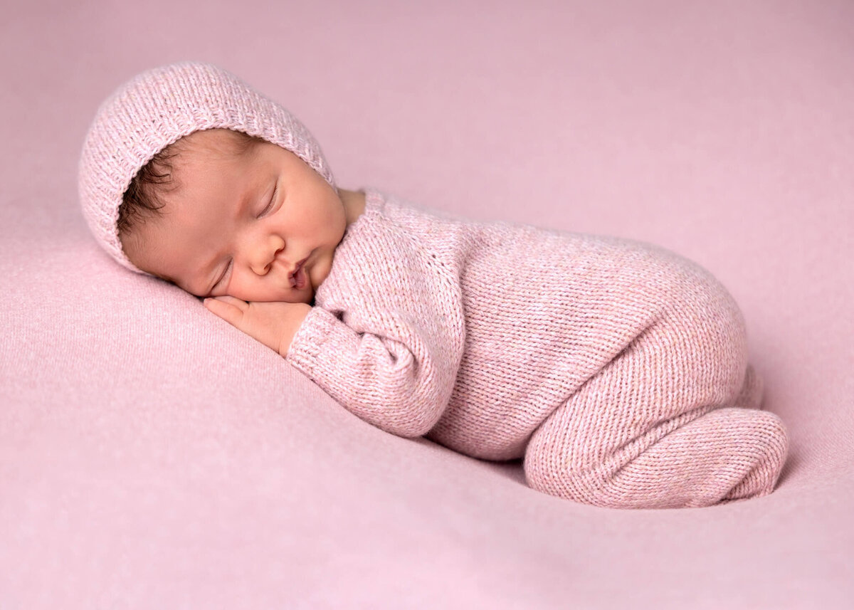 newborn baby wearing a knitted pink romper and bonnet asleep on a pink fabric