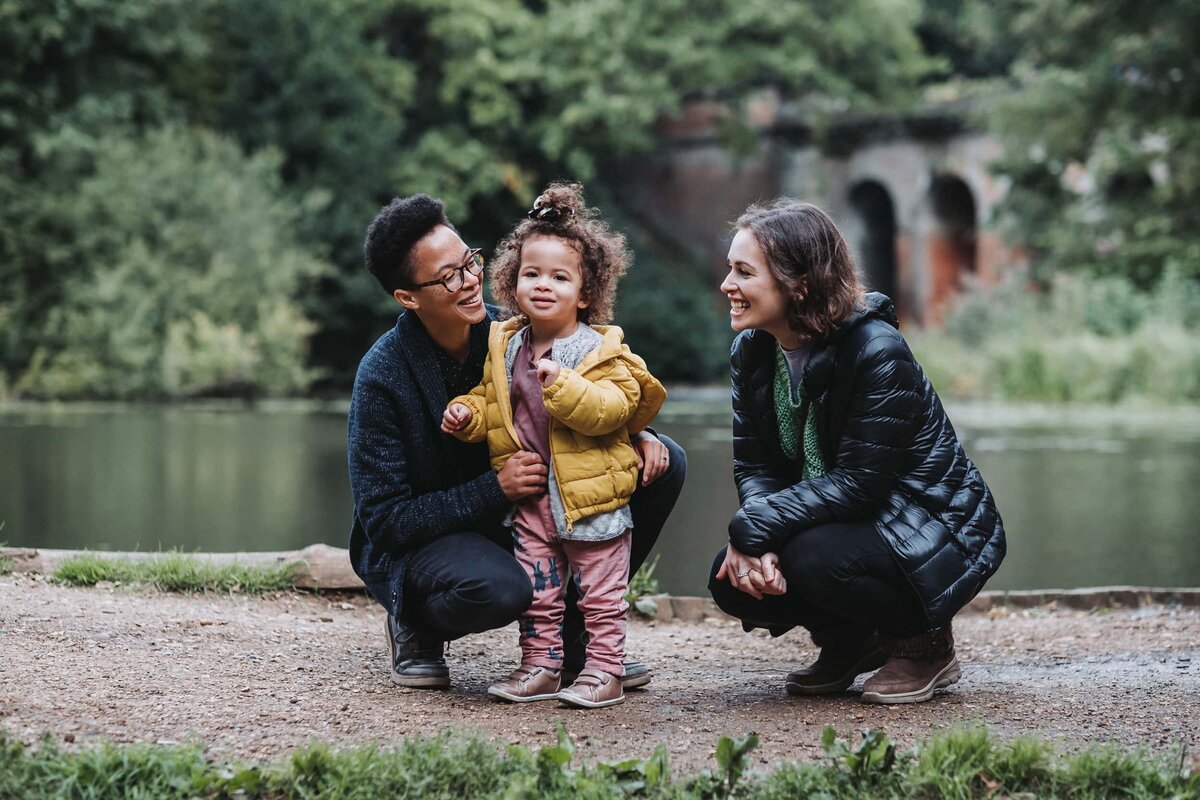 Little girl in pink outfit with yellow coat smiles in front of pond with her mums at Hampstead Heath London