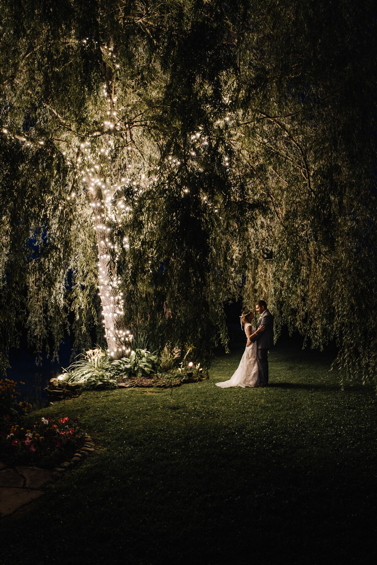 Newlyweds embrace under the lit willow tree at night.