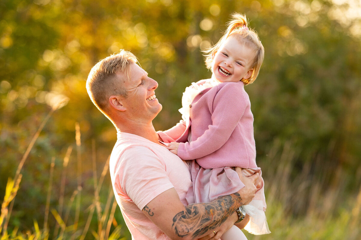 a toddler laughing with her father  in a grassy field during golden hour taken by Ottawa Family Photographer JEMMAN Photography