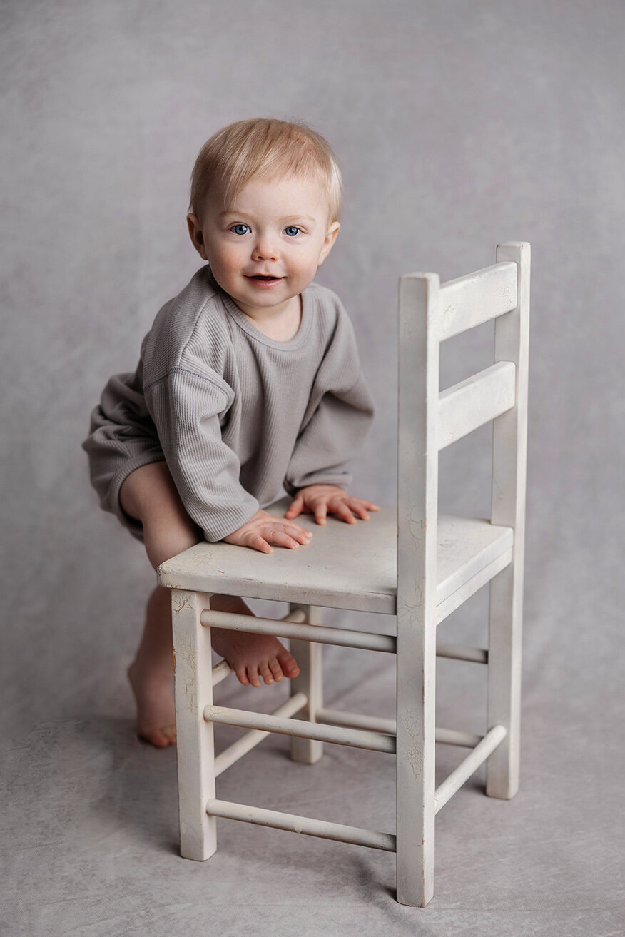 Baby standing next to toddler chair smiling for a neutral tone portrait in Cranberry