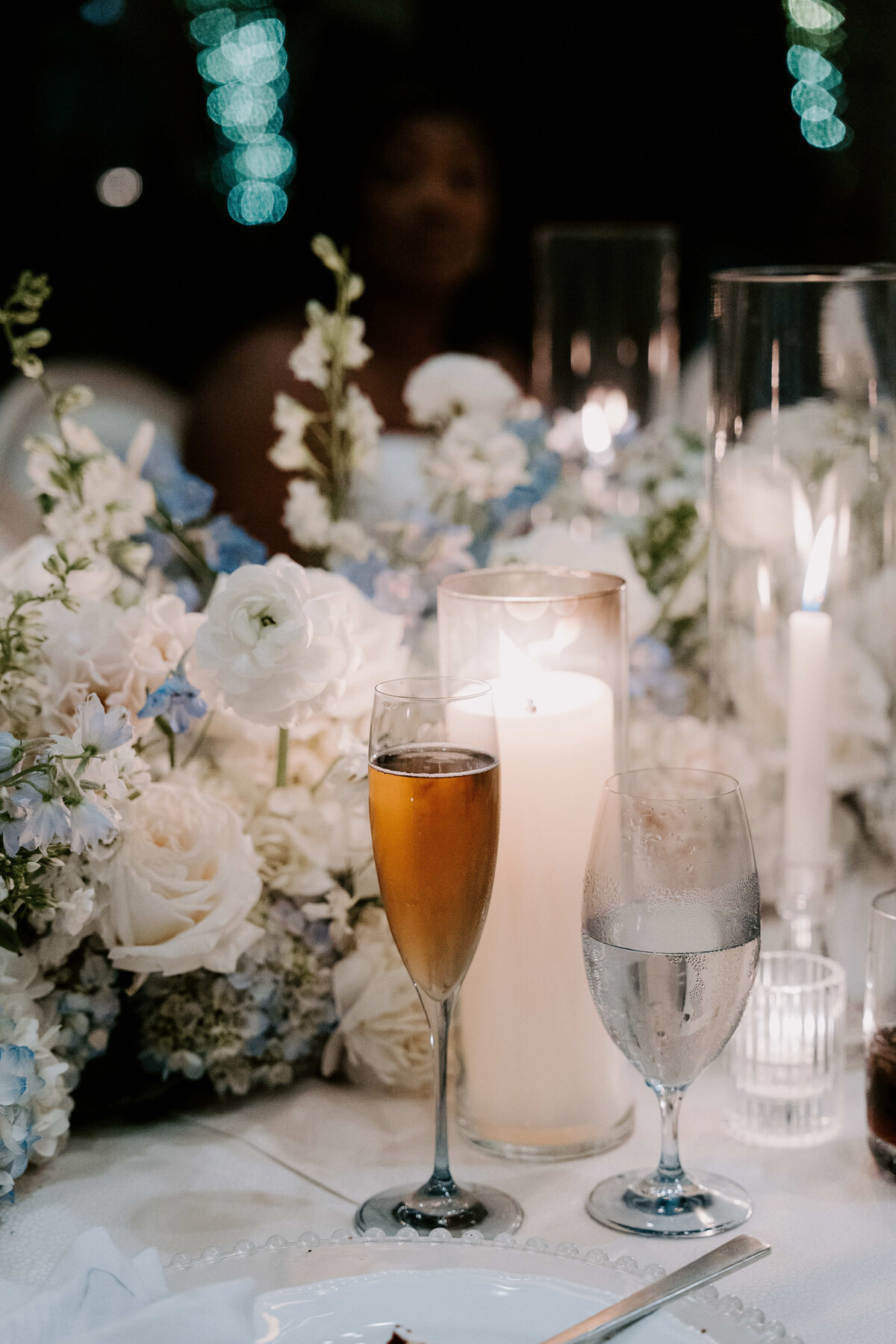 A romantic table setting with soft candlelight, white and light blue flowers, a glass of champagne, and a water glass, creating an elegant and serene atmosphere.