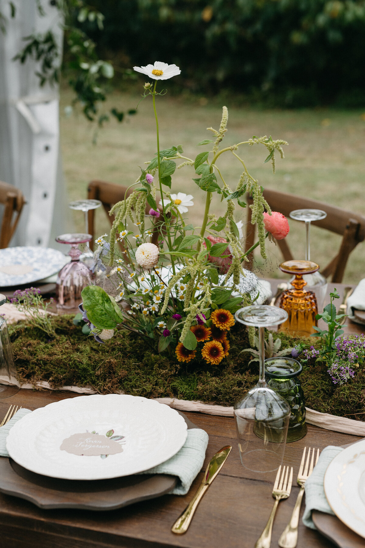 Close-up of a romantic wedding reception table decorated with moss, whimsical wildflowers, taper candles, pastel goblets, and layered floral plates designed by a garden-style wedding florist.