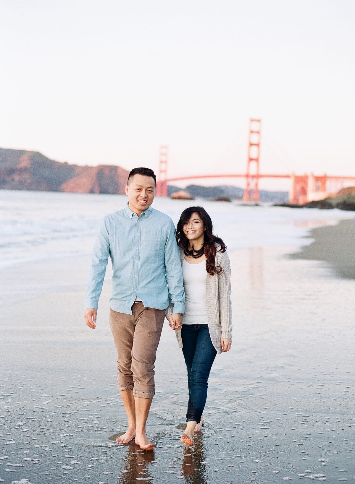 Happy couple hold hands and walk on the beach in front of the Golden Gate Bridge.